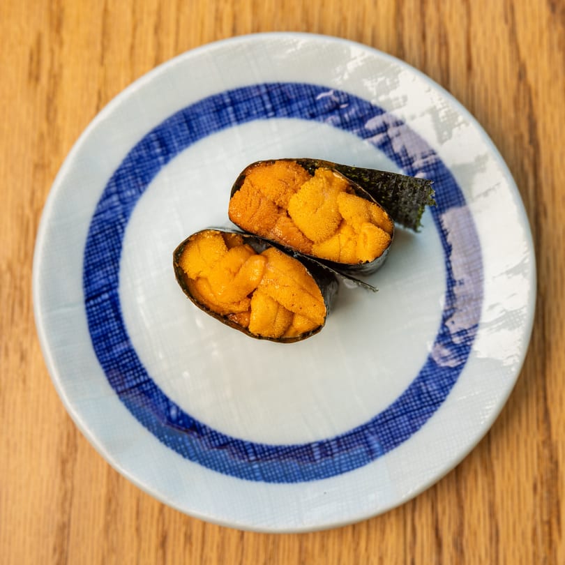 Two pieces of bright orange uni (sea urchin) sushi wrapped in nori on a white and blue plate.