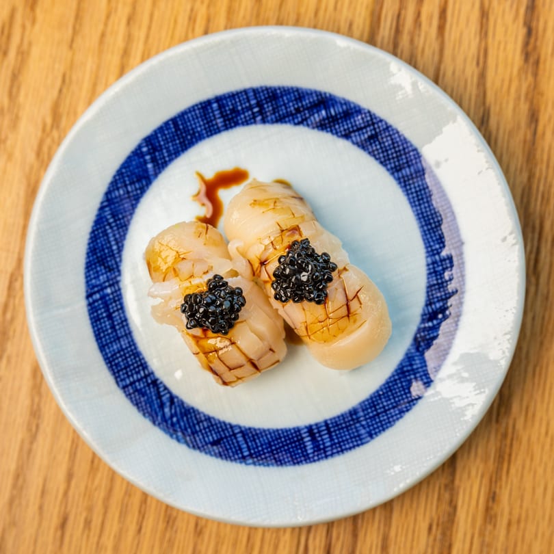 Two pieces of seared scallop nigiri sushi topped with black caviar on a patterned plate.