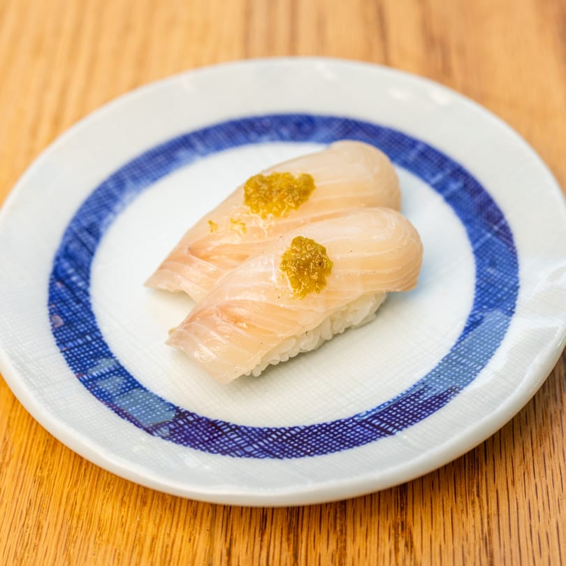 Two pieces of Tatsuki sushi with light fish and green topping on a white and blue plate.